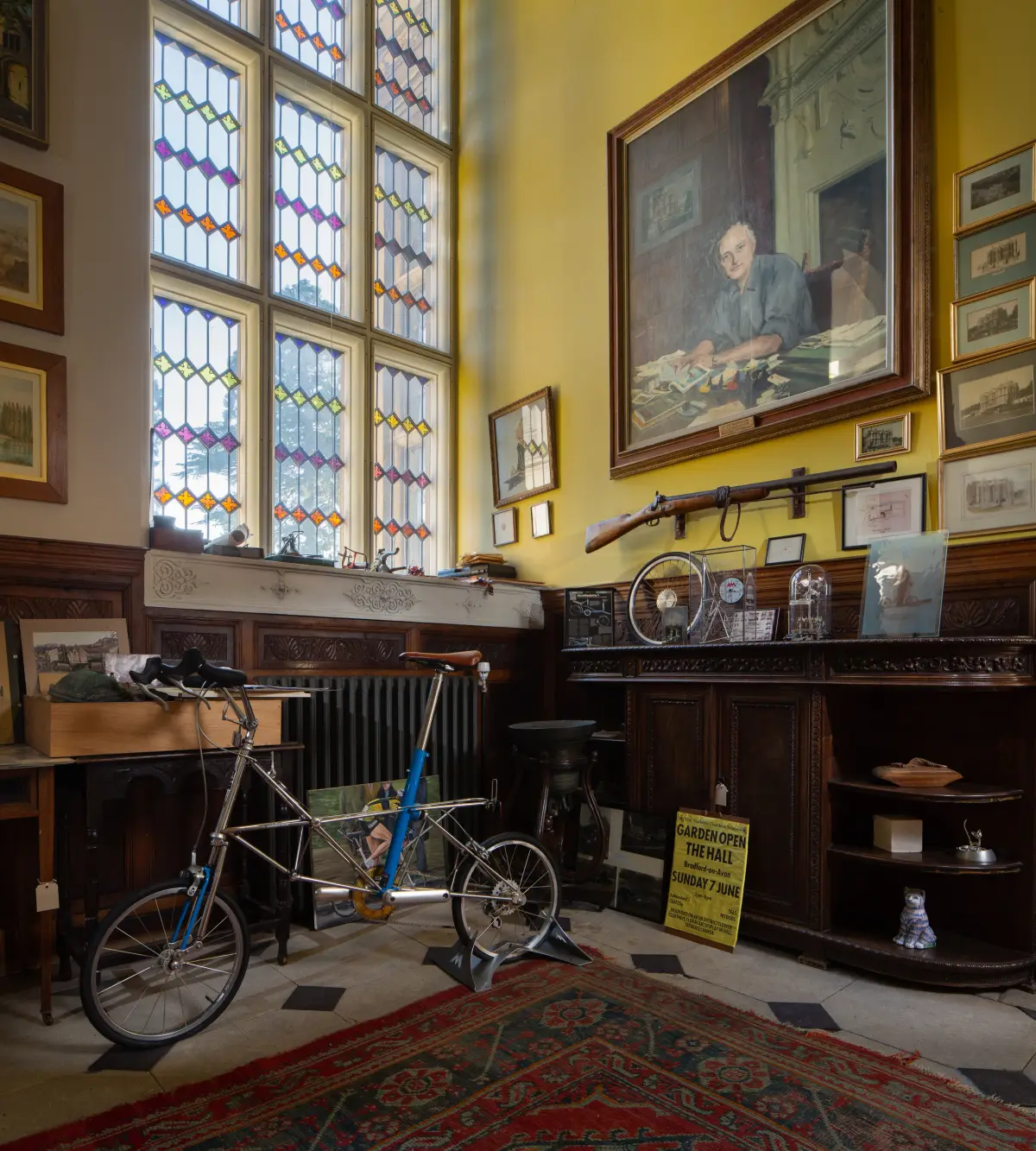 A yellow panelled room with a tall stained glass window, a small-wheeled bicycle on a stand, a portrait of a seated man above the panelling and a rifle mounted on the wall