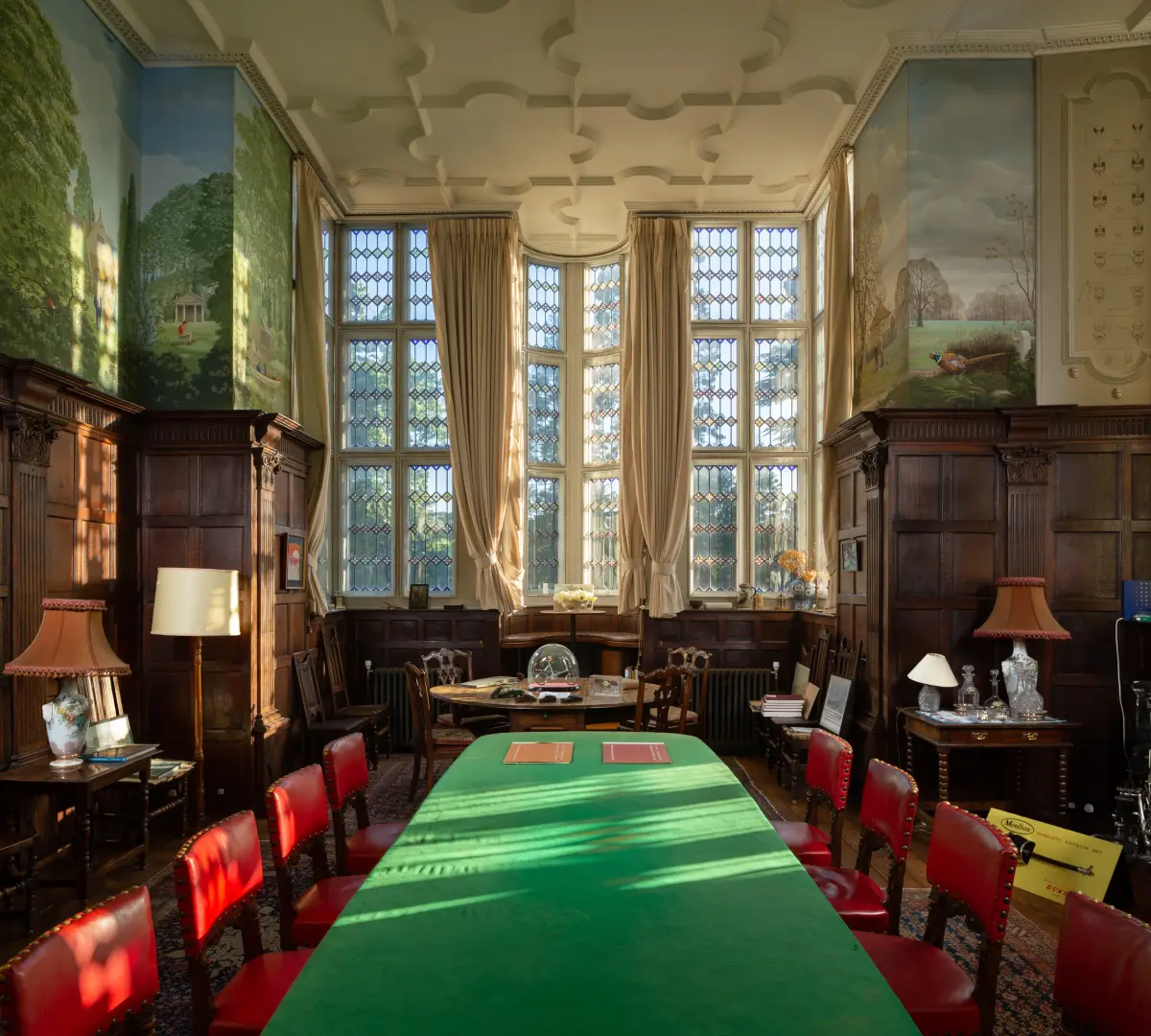 A panelled meeting room under a moulded plaster ceiling, red chairs along a green baize table and painted landscape murals either side of a tall mullioned window