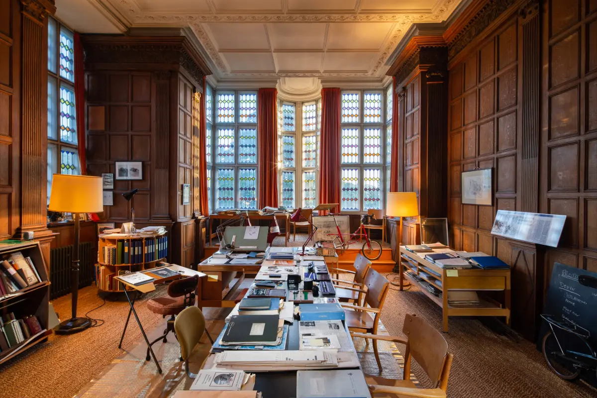 A long panelled library in use as a working archive, tables down the middle stacked with drawings and folders and a bicycle standing at the far end