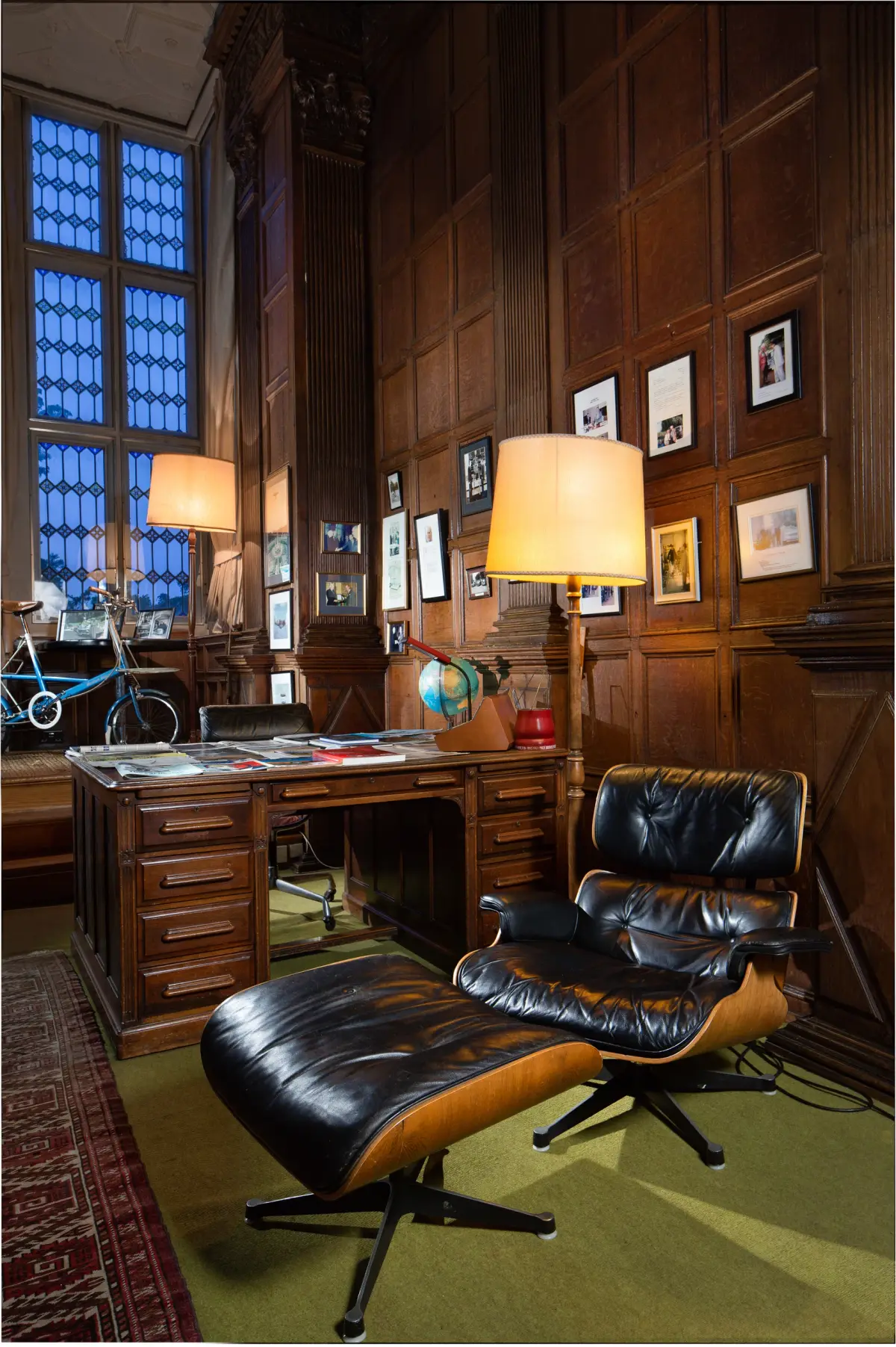 A dark panelled study, a leather lounge chair and footstool beside a pedestal desk under lamplight, framed photographs on the wall behind