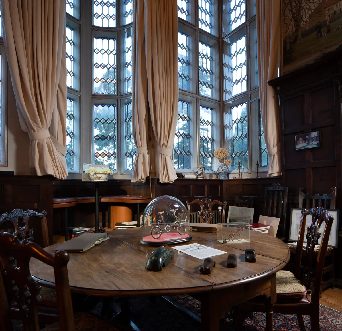 A round dining table below a curtained bay of leaded windows, a bicycle model under a glass dome at its centre