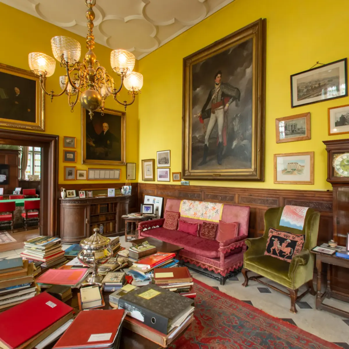 A panorama of the top-lit hall, yellow walls hung with portraits above oak panelling, a brass chandelier overhead and tables of books and archive boxes below