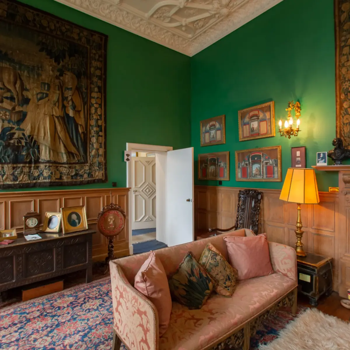 A panorama of the green sitting room, a tapestry over the panelling, a sofa and lamps below a moulded plaster ceiling