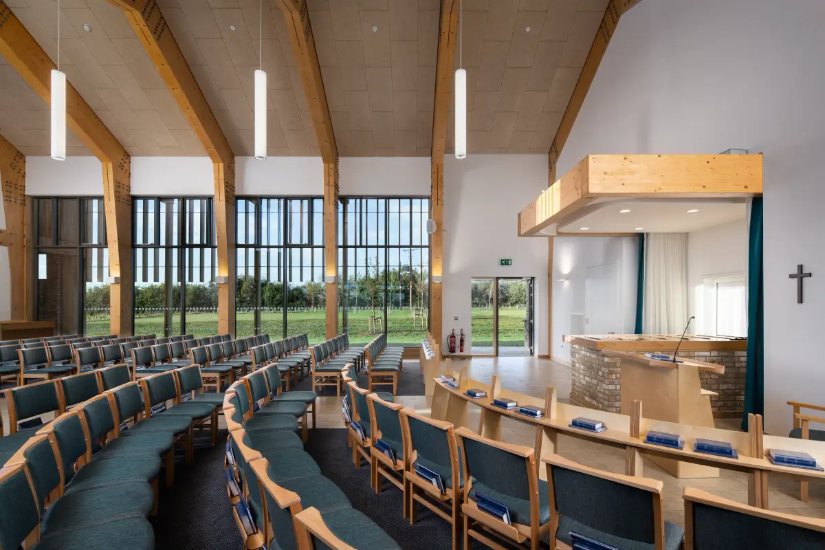The chapel from the side, pendant lights between the trusses, a brick faced lectern and a plain cross on the end wall