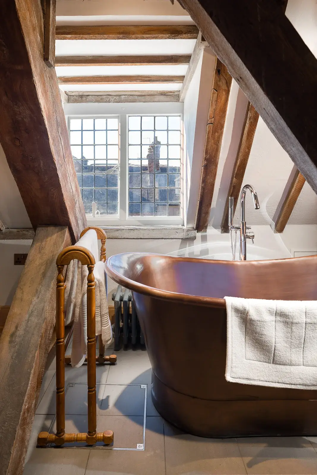 A copper roll top bath under a leaded window in a beamed attic, towel rail and cast iron radiator beside it