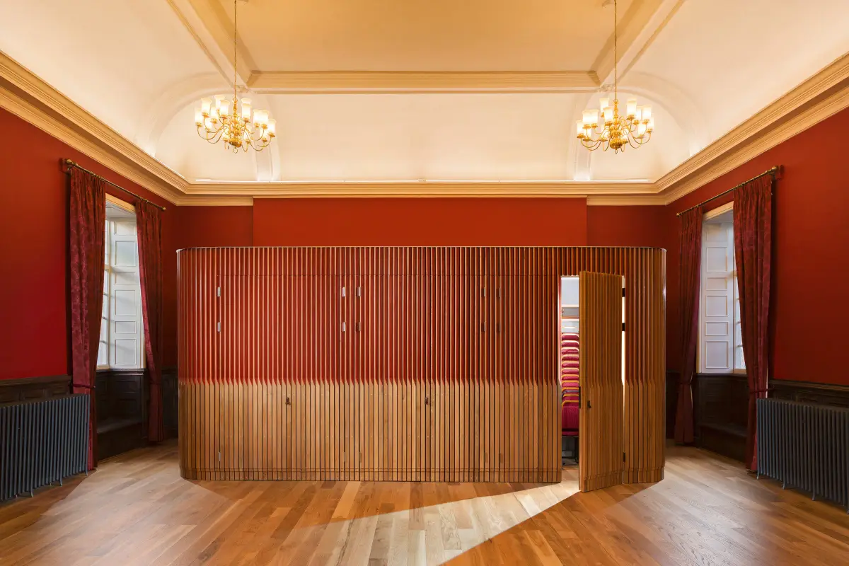 A red walled chamber under two chandeliers, a curved oak slatted pod set into the middle of the floor, one leaf open