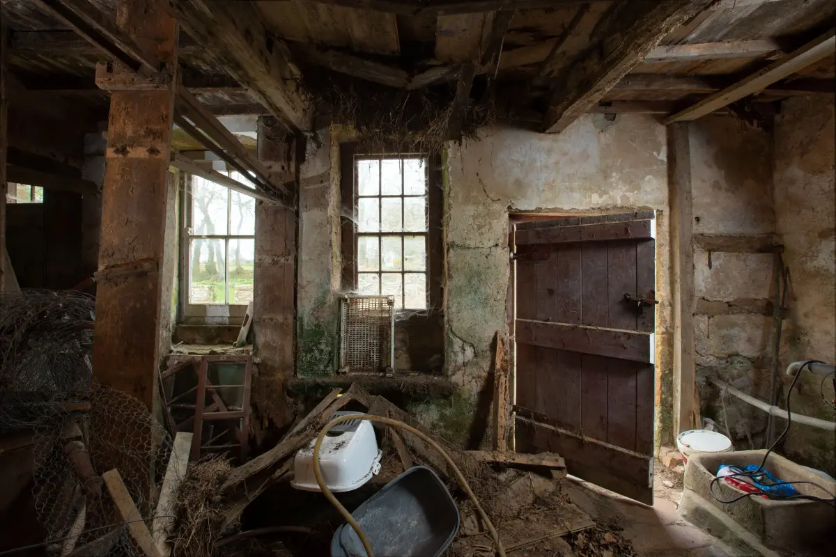 A derelict room stripped back to its timber frame, two small-paned windows, a plank door and debris across the floor