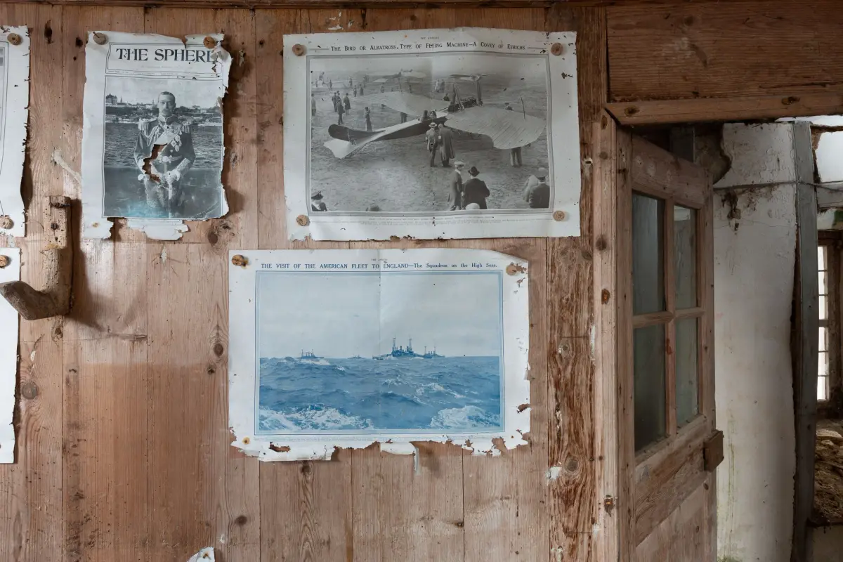 Pages from The Sphere pinned to a plank wall, one showing an early flying machine and another a naval squadron at sea